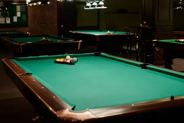 stock image One of several wide wooden tables with rack containing billiards balls on green top standing in front of camera in spacious hall or bar