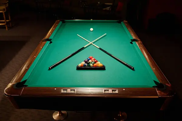 stock image Two crossed billiard cues lying on wide green table between rack with balls and white ball prepared for new leisure game in pub or club