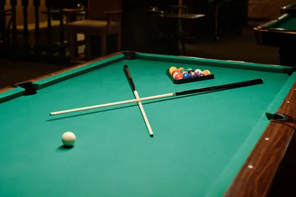 stock image Wide and long table with green top and two crossed billiard cues lying between rack of balls and single white ball ready for game