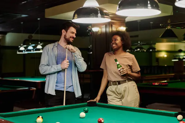 stock image Happy young intercultural couple looking at one another and chatting while standing by green pool table before or after game of billiards