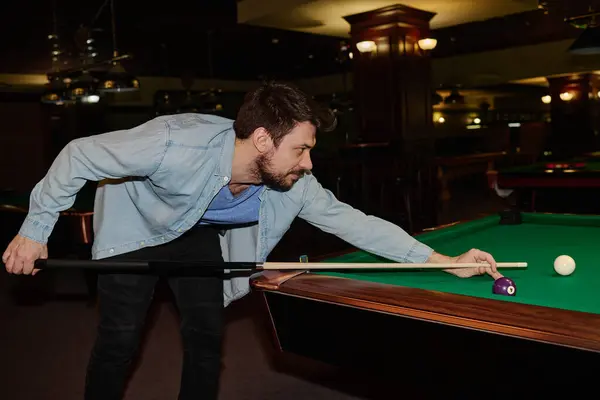 stock image Side view of young man in black jeans and denim shirt standing by wide pool table while hitting one of balls with cue