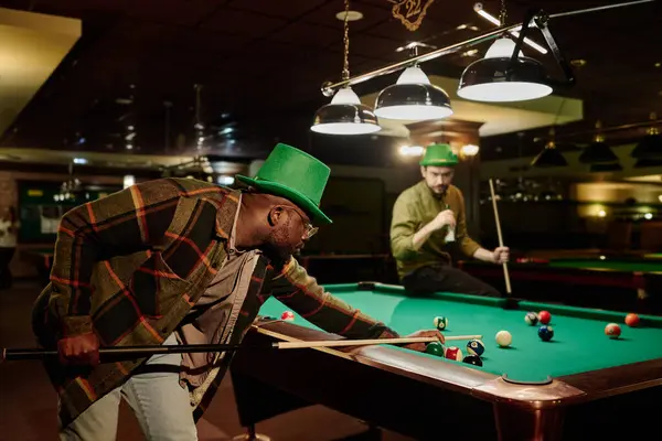 stock image Side view of young African American man in casualwear and green hat hitting pool ball with cue while bending over table in game room