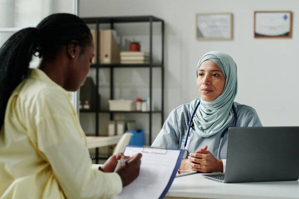 Young serious female doctor in hijab looking at her patient signing medical document or agreement about long term treatment after consultation