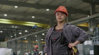 Turuncu şapkalı, tulumlu, kareli gömlekli, belinde poz veren ve fabrika atölyesindeki kameraya bakan İspanyol bir kadının orta yavaş portresi.