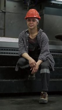 Güvenlik kasketi, iş kıyafeti ve kareli gömlekli kadın fabrika çalışanının dikey görüntüsü. Fabrikada basamaklarda oturuyor ve kameraya bakıyor, boşluğu kopyalıyor.