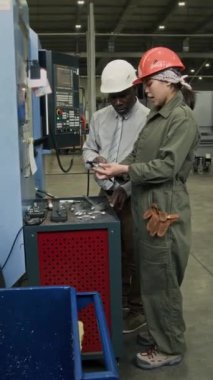 Erkek Afrikalı Amerikalı denetçiye metal parçaları ölçen kadın Latin mühendisin dikey tam yavaşlaması üretim tesisinde üretimin kalitesini kontrol ediyor, başlık takıyor.