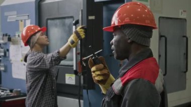 Siyah erkek teknisyenin telsizle fabrika işçileriyle iletişim kurduğu, imalat fabrikasında iş saatleri boyunca emirler verdiği, arka planda çalışan kadın kontrolör.