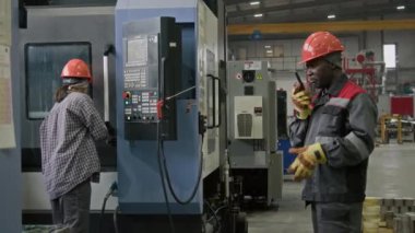Üst düzey siyah ustabaşının koruyucu üniforma giydiği ve fabrika işçileriyle telsiz aracılığıyla iletişim kurduğu prodüksiyon sürecini kontrol ettiği arka planda çalışan bir işçinin orta boy fotoğrafı.