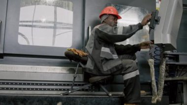 Siyah erkek teknisyenlerin koruyucu tulumları ve baret giyip endüstriyel kontrolörün yanında otururken ve fabrikada otomatikleştirilmiş makine üretim sürecini yönetirken tam pozu.