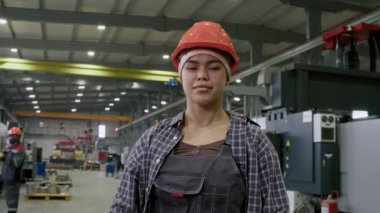 Portakal rengi güvenlik kasketi, iş kıyafetleri ve damalı gömlekli Latin işçilerin orta yavaş kahraman portresi. Kameraya güvenle bakıyor ve üretim tesisinde iş günü boyunca kollarını kavuşturuyor.