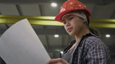Düşük açılı, turuncu şapkalı İspanyol ustabaşı makine üretim fabrikasındaki resim makinesi montaj planına bakıyor.