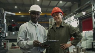 Formalite icabı giyinmiş yetişkin bir işadamının orta boy portresi, pano ile birlikte üniformalı kadın Latin fabrika işçisi ile poz veriyor, atölyede kameraya bakıyor, ikisi de şapka takıyor.