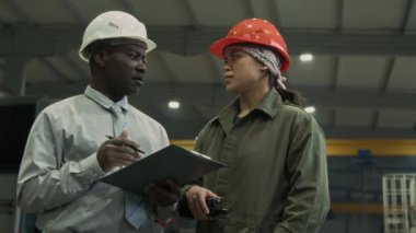 Üst düzey Afro-Amerikan iş adamının ofis giyimi ve beyaz şapkalı ve pano ile İspanyol fabrika mühendisi kadınla konuşurken orta yavaş çekimi, atölyede üretim sürecini tartışırken.