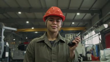 Portakal rengi şapkalı, tulumlu, fabrika atölyesinde elinde telsizle kameraya bakarak poz veren Latin kökenli genç kadının yavaş yavaş portresi.
