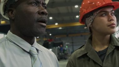 Formalite icabı iş adamlarının yakın çekimleri ve kadın fabrika müdürünün telsize konuşması, ikisi de sert şapkalı, fabrika atölyesindeki üretimin kalitesini kontrol ediyor.