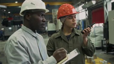 Tişörtlü ve kravatlı olgun erkek Afrikalı Amerikalı denetçinin orta yavaşlığında rapor yazarken Latin ustabaşı telsizle emir veriyor, üretimin kalitesini kontrol ediyor.