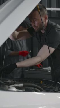 Asyalı meslektaşı alet çantasında İngiliz anahtarı ararken, beyaz tenli erkek araba tamircisinin dipstick 'i çıkardığı ve motor yağ seviyesini ölçtüğü orta boy dikey çekim.