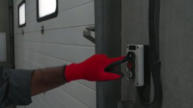 Yetişkin Asyalı araba tamircisinin gri tulumlu ve kırmızı eldivenli orta boy yakın çekim görüntüsü. Garaj kapısını açmak için kapı düğmesine basarken, sabah vardiyasında oto tamirhanesi açılışı.