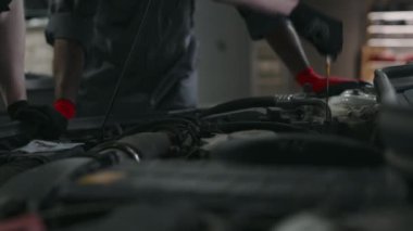 Gri tulumlu iki erkek araba tamircisinin, garajda müşteri arabasına servis yaparken, motor yağı seviyesini kontrol ederken ve dokuyla sondayı silerken orta boy yakın çekim görüntüleri.