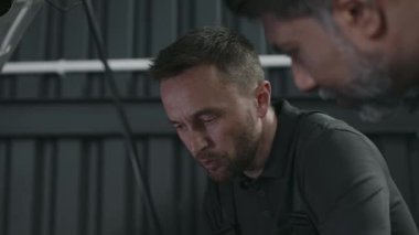 Asyalı iş arkadaşıyla araba kaputunun yanında duran beyaz erkek araba tamircisinin orta ölçekli yakın çekimi sırasında motor bileşenlerini kontrol ederken planlanan hizmet sırasında arıza nedeni aranıyor.