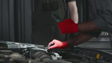 Gri tulumlu ve kırmızı eldivenli orta yaşlı Hintli teknisyenin voltmetre ile araba motorundaki voltajı kontrol ederken araba garajında bakım yaparken orta boy yakın çekim çekimi.