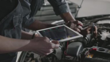Kadın teknisyen elinde tablet bilgisayar zeka tanısı yazılımı tutarken, Asyalı erkek tamirci, amperemeter ile voltajı kontrol ederken, garajda arabayı tamir ederken...