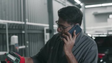 Olgun Asyalı tamircinin el feneri denetimi ve araba süspansiyonuyla orta boy fotoğrafı müşteriyle cep telefonunda konuşurken, oto servis istasyonunda tamirat sırasında.