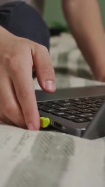 Beyaz çocuğun yatağında oturup, elektronik işitme cihazını veri kablosuyla birbirine bağlayıp, müzik dinlemek ya da online kolej dersleri dinlemek için kulağının arkasına koyduğu dikey eğik görüntü.