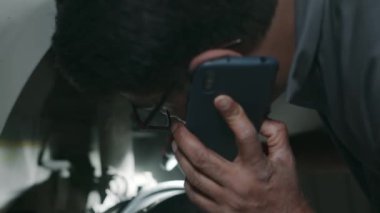 Erkek garaj teknisyeninin arabadaki süspansiyon ve direksiyon mekanizmasını kontrol ederken müşteriyle cep telefonundaki bakım işini tartışırken yakın plan fotoğrafı.