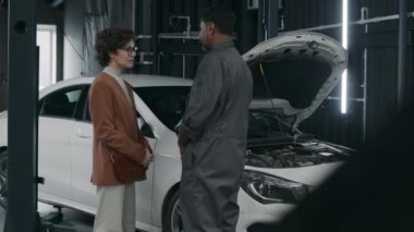 Tamirden sonra garajdan araba toplayan beyaz bir kadın sürücü. Tekerlekleri ve motoru işaret eden Hintli tamirci. Onarımları tarif eden kadın.
