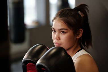 Esmer kız boks eldivenlerini gözünün önünde tutuyor ve modern spor salonunda antrenman yaparken kum torbasına bakıyor.