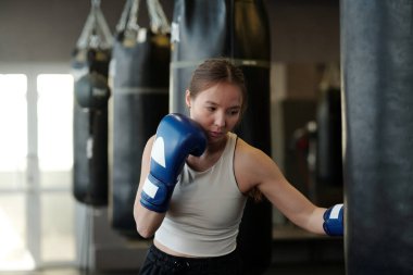 Beyaz tişörtlü ve boks eldivenli genç aktif bir kadın modern spor salonunda kum torbasının önünde dururken temel tekmeleri deniyor.