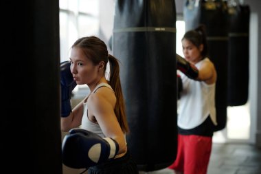 Boks eldivenli genç sporcu boks yaparken kum torbaları arasında duruyor ve diğer boksörlere karşı temel egzersiz ve tekme yapıyor.