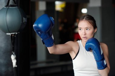 Boks eldivenli genç aktif bayan sporcu spor salonunda antrenman yaparken kum torbasının önünde tekme ve savunma egzersizleri yapıyor.