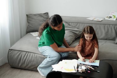 Küçük bir masada küçük bir kanepede oturmuş pastel boyalarla resim çizerken genç bir bakıcı ona yardım ediyor.