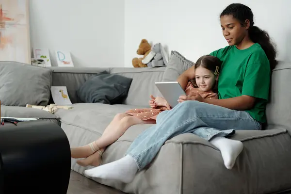 stock image Two happy intercultural girls in casual attire watching online cartoons or curious video while both sitting on comfortable couch at home