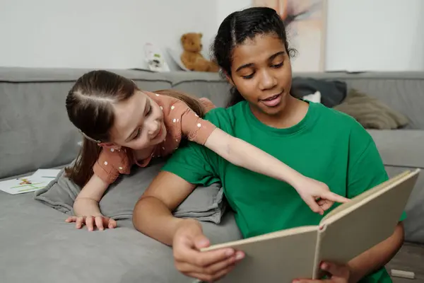 stock image Happy little girl relaxing on sofa and pointing at page of open book with fairy tales or stories for kids while talking to teenage sitter