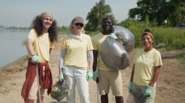 Sarı tişörtlü, lastik eldivenli, kumsalda çöp torbaları üstüne çöp torbalarıyla yan yana duran ve kameraya bakıp gülümseyen orta karar gönüllü takım.