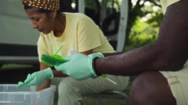 Orta boy İspanyol kadın ve Afro-Amerikan erkeğin açık havada çöp toplama etkinliğinde taburelerde oturup tek kullanımlık plastik çatal bıçakları renkli kaplara ayırdığı bir fotoğraf.