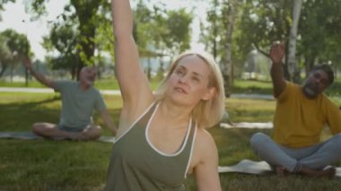 Güneşli bir yaz günü parkta, açık hava grubu eğitimi sırasında beyaz kadın spor eğitmeninin, çeşitli yaşlılarla yan yana eğildiği orta boy bir fotoğraf.