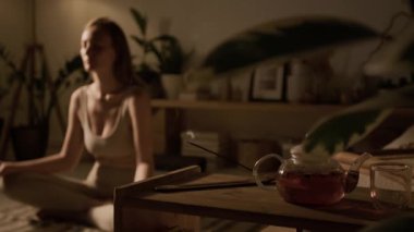 Spor giyim kuşağı giymiş bir kadının bacak bacak bacak üstüne atmış, tütsü yakmaya odaklanmış, evinde bitki çayıyla cam çaydanlığın yanındaki masada tütsü yaktığı bir fotoğraf.