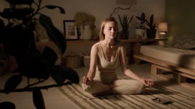 Sukhasana pozu vermiş bej renkli aktif giysili genç beyaz bir kadın. Bacakları çaprazlanmış, meditasyon yapıyor, gözleri kapalı, ellerinde Gyan Mudra, evinde hafif bir ışık.