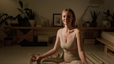 Tam portre halinde bej bluzlu beyaz yogalı gülümseme ve sukhasana pozisyonunda yerde oturan taytlar. Elleri Gyan Mudra 'da ve kameraya bakıyor, rahat atmosfer, yumuşak ışık.