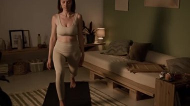 Orta yaşlı, beyaz tenli, bej bluzlu ve taytlı bir kadın akşamları süslü, rahat bir odada siyah minder üzerinde yoga yapıyor, savaşçı pozu alıyor ya da virabhadrasana yapıyor ve üstte namaste yapıyor.