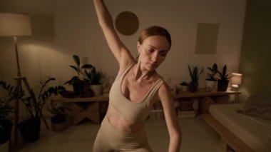 Orta yaşlı, bej bluzlu, tayt giymiş, evde yoga yapan, kenar açılı poz veren ya da utthita parsvakonasana yapan, dekore edilmiş ve rahat bir odada uzanan beyaz bir kadın.