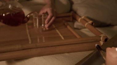 Evdeki sıcak atmosferik çay seremonisi sırasında el kamerasında yanan mum ve yüzsüz kadının camdan çaydanlığa bitkisel çay doldurması, yumuşak ışık.