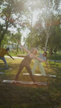 Yaşlılar için yoga eğitimi veren, güneşli yaz gününde şehir parkında utthita trikonasana yapan orta yaşlı bir kadının el kamerası görüntüsü.