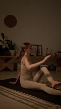 Dikey çekimde bej bluzlu beyaz kadın ve taytlı kadın rahat bir odada yin yoga yapıyor ve omurga büküyor ya da ardha matsyendrasana yapıyor.