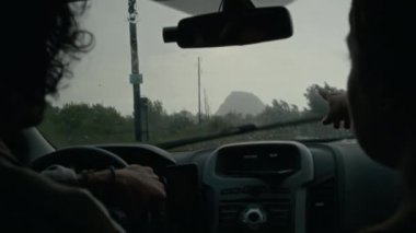 Genç beyaz bir erkek ve kadının yağmurda birlikte araba sürerken, ıssız bir yerde, kırsal alanda, toprak yolda, dağlara uzaklığı gösteren bir kızın orta boy arka çekimi.