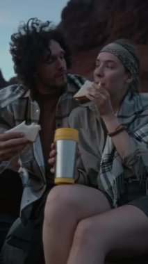 Genç beyaz çiftin birlikte pikapta otururken, akşam yemeğinde çayla sandviç yerken çölde manzaralı günbatımını seyrederken orta boy dikey çekimleri.